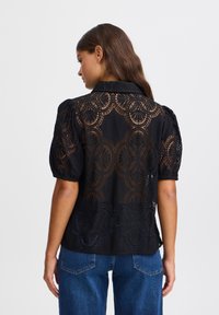 Blusa nera con maniche a sbuffo, caratterizzata da un intricato motivo in pizzo e un design con ritagli. Indossata con jeans blu a vita alta. Nessun marchio visibile.