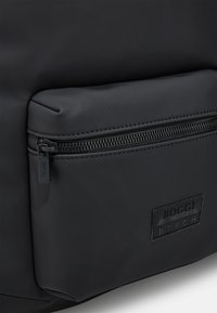 Mochila negra con un acabado suave y mate, que presenta un bolsillo frontal con cremallera, cremallera negra y un parche de marca texturizado.