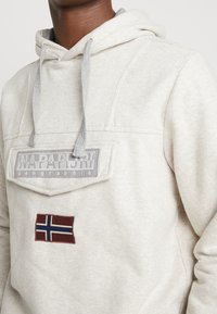 Sudadera gris claro con cordones a juego. Presenta un gran parche con el logo y una bandera noruega bordada en el pecho. Textura de tela suave.