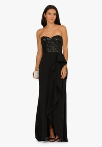 Czarna suknia z bezrękawnikowym gorsetem ozdobionym cekinami i zwiewną spódnicą z falbaną. Metaliczna kopertówka i wysokie obcasy dopełniają stylizację.