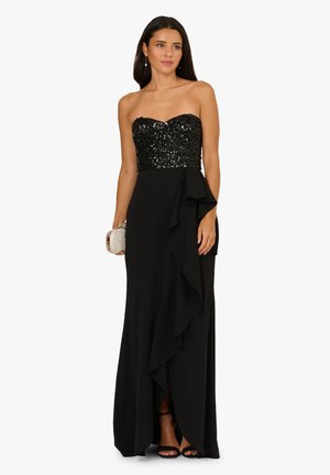 Schwarzes Kleid mit einem glitzernden, trägerlosen Oberteil und einem fließenden Rock mit Rüschendetail. Eine metallische Clutch und hohe Absätze ergänzen das Outfit.