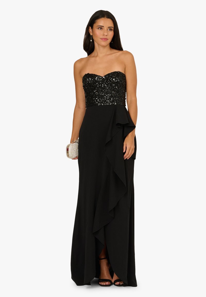 Czarna suknia z bezrękawnikowym gorsetem ozdobionym cekinami i zwiewną spódnicą z falbaną. Metaliczna kopertówka i wysokie obcasy dopełniają stylizację.