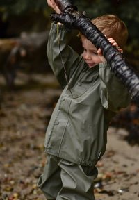 Traje de lluvia verde oliva hecho de material impermeable con botones a presión, cintura elástica y mangas largas. Niño sostiene una rama negra.