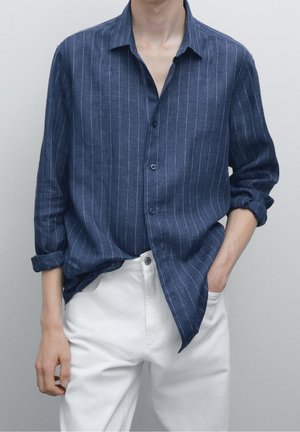 Homme portant une chemise bleu marine à rayures verticales et manches longues retroussées, avec un pantalon blanc, une main dans la poche, sur un fond uni.