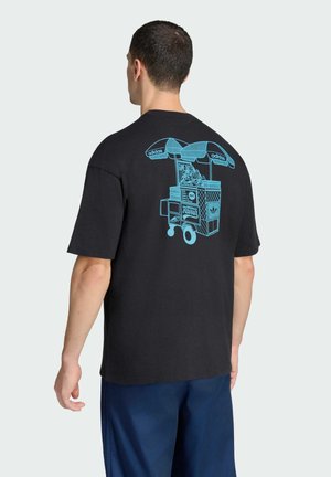 Uomo che indossa una maglietta nera con una grafica blu di un carrello di street food con etichetta Adidas e Hawaii sulla schiena, in piedi di spalle.