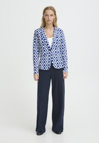 Blazer avec un motif géométrique bleu et blanc, fermeture à un bouton, associé à un pantalon ajusté bleu marine, texture lisse et design structuré.