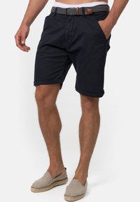 Shorts en coton bleu marine avec des ourlets retroussés, avec une ceinture tissée à motifs et des poches latérales. Portés avec des espadrilles gris clair.