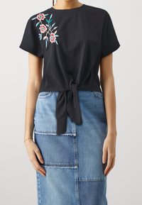 Zwarte katoenen top met korte mouwen, een strikdetail aan de voorkant en bloemrijke borduursels in roze en blauw. Gecombineerd met een patchwork denim rok.