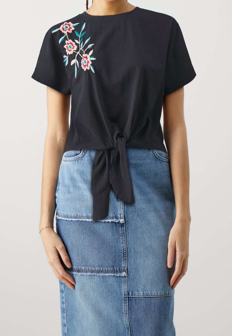 Zwarte katoenen top met korte mouwen, een strikdetail aan de voorkant en bloemrijke borduursels in roze en blauw. Gecombineerd met een patchwork denim rok.