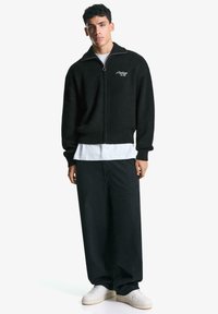 Black ribbed zip-up sweater with a high collar, paired with loose black pants and white sneakers, featuring a logo on the chest.