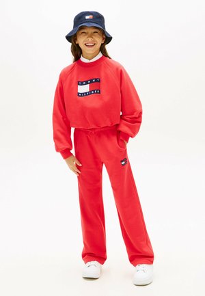 Joven sonriente, vistiendo sudadera y pantalones rojos de Tommy Hilfiger, zapatillas blancas y un sombrero tipo bucket azul marino.