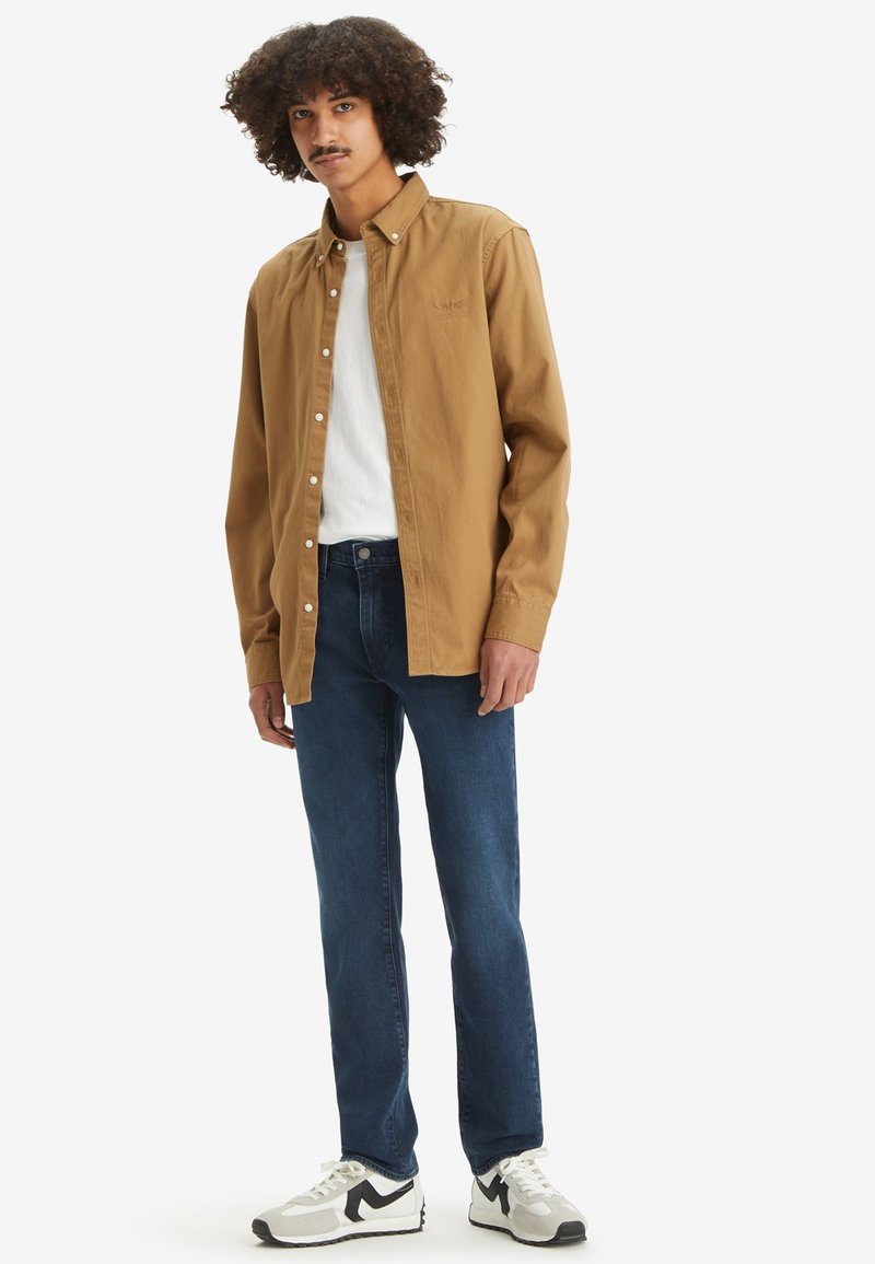 Chemise en coton marron à boutons sur un T-shirt blanc, associée à un jean bleu et des baskets noir et blanc. Le mannequin a les cheveux bouclés.