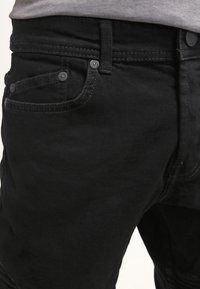 Jeans de mezclilla negra con un diseño clásico de cinco bolsillos, costuras detalladas y botones de metal. La tela tiene un acabado texturizado.