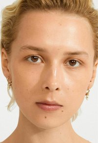 Nahaufnahme einer Person mit heller Haut, kurzen blonden Haaren, braunen Augen, die kleine goldene Creolenohrringe mit Charms trägt.