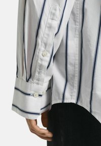 Witte shirt met marineblauwe verticale strepen, knoopmanchetten en GANT-logo borduursel op de mouw. De stof lijkt glad en licht van gewicht.
