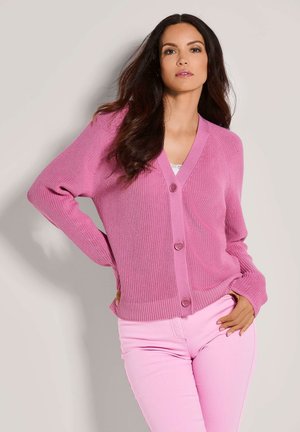 Vrouw met lang donker haar, gekleed in een roze gebotoneerde cardigan en lichtroze broek, staand met één hand op haar heup tegen een lichte achtergrond.