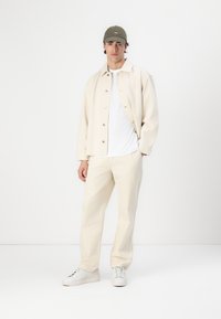 Beige knap-ned jakke med lange ærmer, hvid t-shirt, beige bukser og hvide sneakers; toppet med en grøn baseball cap. Minimalistisk design.