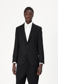 Costume noir à deux boutons avec revers crantés, coupe ajustée et poches latérales, associé à une chemise habillée blanche. Texture de tissu lisse.