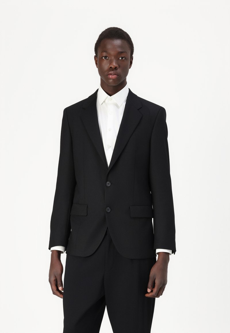 Abito nero a due bottoni con rever a punta, slim fit e tasche laterali, abbinato a una camicia bianca. Tessuto dalla texture liscia.