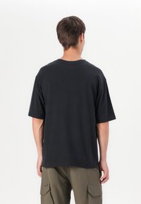 Personne portant un t-shirt noir uni à manches courtes et un pantalon cargo vert olive, de dos sur un fond blanc uni.