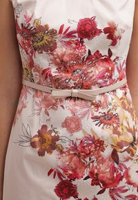 Robe fleurie avec des fleurs roses et rouges sur un fond clair, dotée d'une texture lisse, d'une coupe ajustée et d'une ceinture nude assortie ornée d'un nœud.