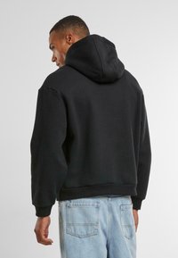 Felpa nera con cappuccio regolabile, in cotone, vestibilità rilassata, polsini e orlo a coste. Indossata con jeans azzurri chiari, vista da dietro.