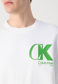 Fehér pulóver bordás nyakkal, nagy zöld "CK" logóval és "Calvin Klein EST - 1968" felirattal az elején.