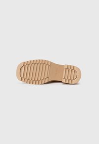 Talpă de pantof de cauciuc de nuanță tan, cu textură striată, având un logo embosat. Finisaj neted și fără detalii vizibile de model sau hardware.