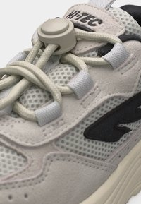 Közelkép egy bézs sportcipőről, amely hálós anyagból készült, velúr ráhajtásokkal, fekete logóval és rugalmas fűzőkkel, amelyeket kerek műanyag csúszkával rögzítenek.