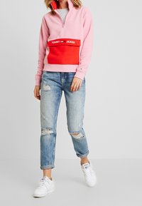 Różowy polarowy sweter z pół zamkiem błyskawicznym i czerwoną kieszenią z przodu, z logo "TOMMY JEANS"; w połączeniu z podartych jasnoniebieskich dżinsami i białymi trampkami.