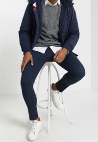 Laivastonsininen puffatakki punaisilla ja valkoisilla yksityiskohdilla, harmaa neulepaita, valkoinen kauluspaita, laivastonsiniset housut ja valkoiset lenkkitossut. Malli istuu jakkaralla.