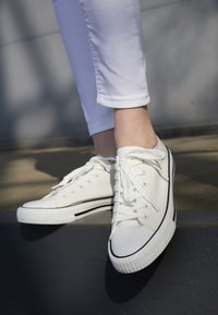 White canvas sneakers with black lines, round toe, and rubber soles. Laces are tied. Worn with rolled-up light-colored pants.
