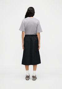 Γυναίκα με μακριά σκοτεινά μαλλιά στέκεται με την πλάτη, φοράει γκρι κοντομάνικο μπλουζάκι, μαύρη φούστα μέχρι τα γόνατα, λευκές κάλτσες και γκρι αθλητικά παπούτσια.