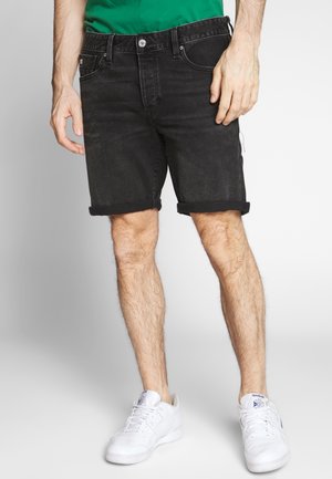 Mann trägt hochgekrempelte schwarze Jeansshorts mit grünem Hemd und weißen Sneakers und steht vor einem einfarbigen Hintergrund.