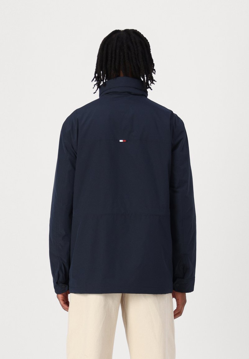 Marineblaue wasserdichte Jacke mit hohem Kragen, ausgestattet mit einem kleinen rot-weiß-blauen Logo-Detail auf der Rückseite. Leicht und strapazierfähig.