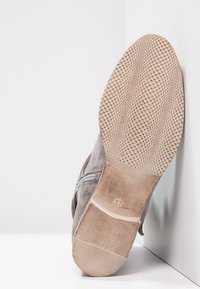 Zapato de ante gris con una suela de goma texturizada. La parte inferior presenta pequeñas agarraderas circulares y una etiqueta de tamaño, mostrando áreas lisas y ligeramente desgastadas.