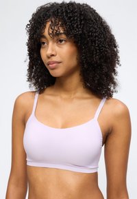 Mladá žena s kudrnatými vlasy, která má na sobě světle fialovou bezešvou podprsenku, mírně otočená nalevo na jednobarevném pozadí.