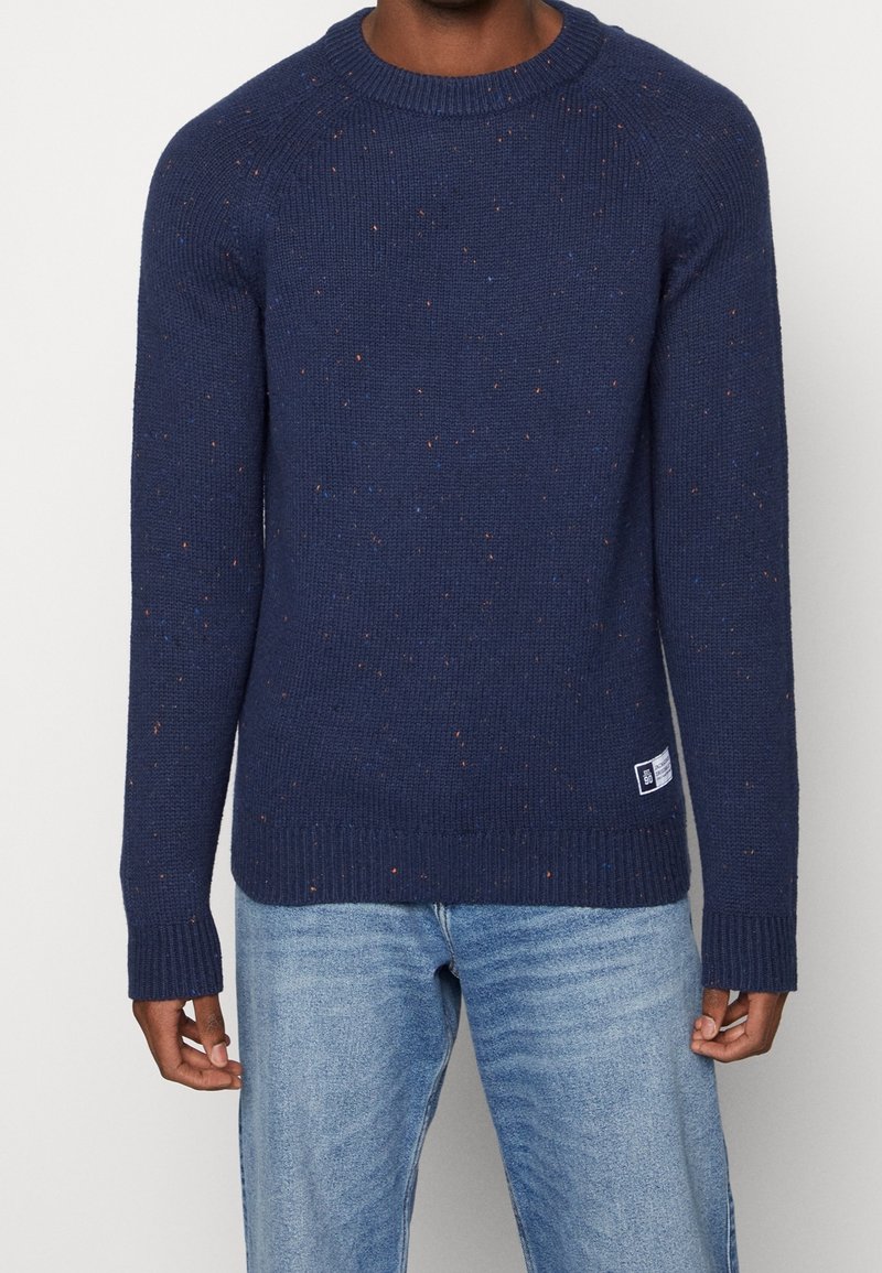 Man med en marinblå prickig stickad tröja och ljusblå jeans, stående mot en enkel vit bakgrund.