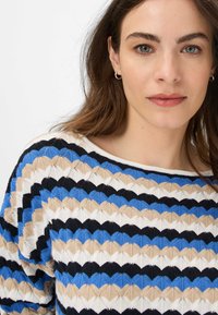 Strickpullover mit weitem Halsausschnitt, der ein Zickzackmuster in Blau, Beige, Schwarz und Weiß zeigt. Textil mit strukturierter Oberfläche und lockerer Passform.