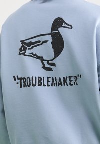 Sudadera azul claro con un gráfico de un pato negro y la palabra "TROUBLEMAKER" impresa debajo en una fuente negrita y desgastada.