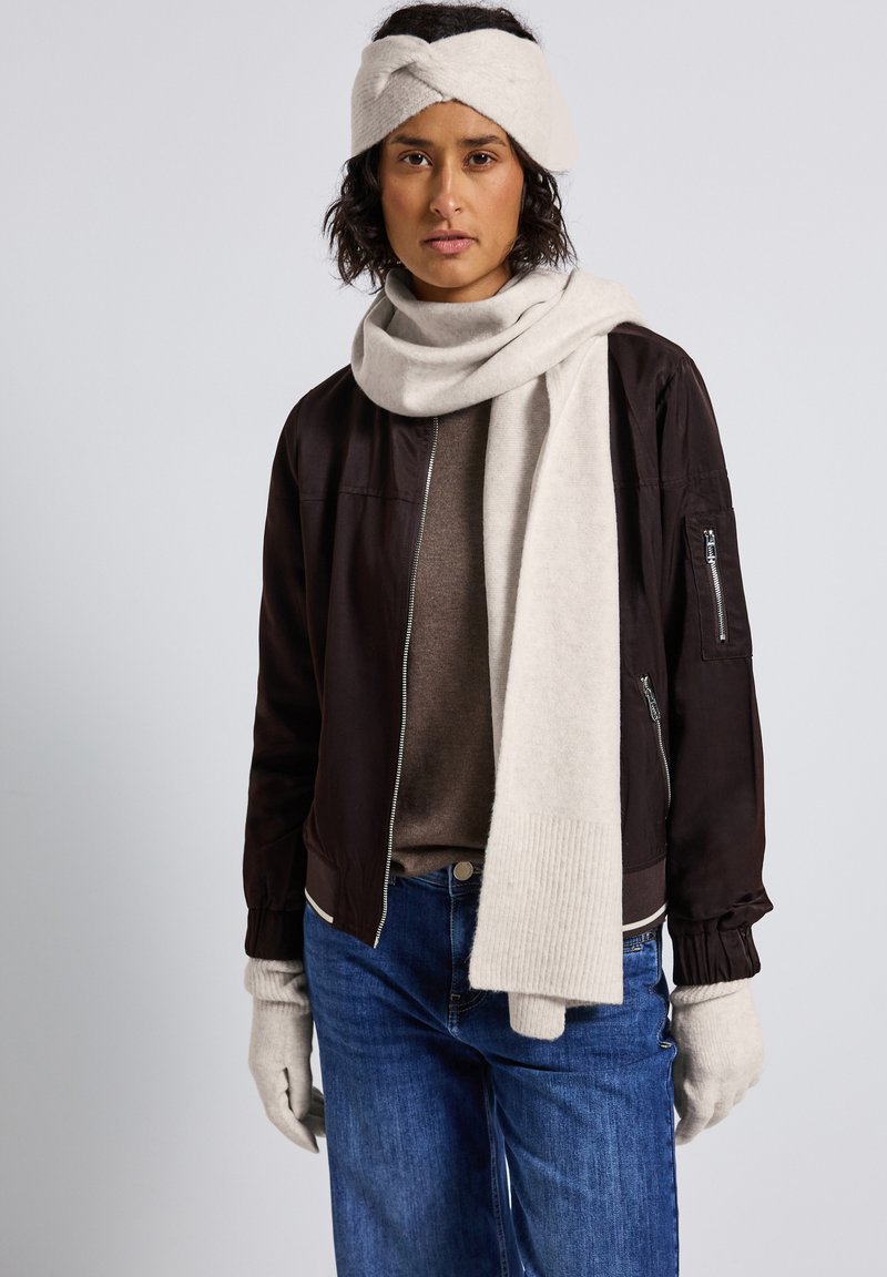 Mørkebrun bomberjakke, beige tørklæde, matchende strikhovedbånd, tan sweater, blå jeans og lyse handsker. Bløde teksturer og neutrale farver.