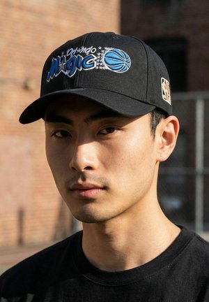 Jeune homme portant une casquette de baseball noire Orlando Magic et une chemise noire, debout en plein air près d'un mur en briques à la lumière du jour.