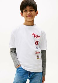 T-shirt bianca in cotone con testo ricamato rosso e patch. Design a girocollo, maniche corte, indossata sopra una camicia a maniche lunghe grigia. Jeans blu.