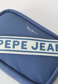 Modra tkaninska torbica z zaobljenimi robovi, opremljena s kremno naramnico z napisom "PEPE JEANS" v modrem besedilu in majhnim logotipom na površini.