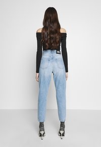 Kvinna med långt brunt hår som bär en off-shoulder svart topp, ljusblå jeans med hög midja, mönstrade strumpor och silvriga högklackade skor, vänder sig bort.