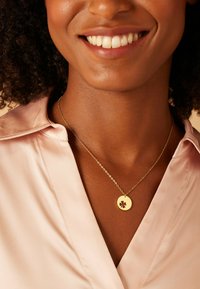 Collier en or avec un pendentif circulaire présentant un design de fleur découpée, porté sur une chemise en satin rose clair.
