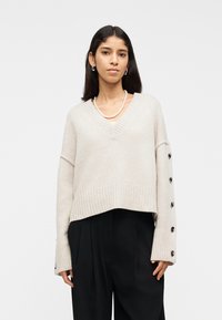 Femme portant un pull à col en V beige avec des boutons sur les manches, un pantalon noir, un collier de perles et des boucles d'oreilles argentées en forme de créole, sur un fond blanc.