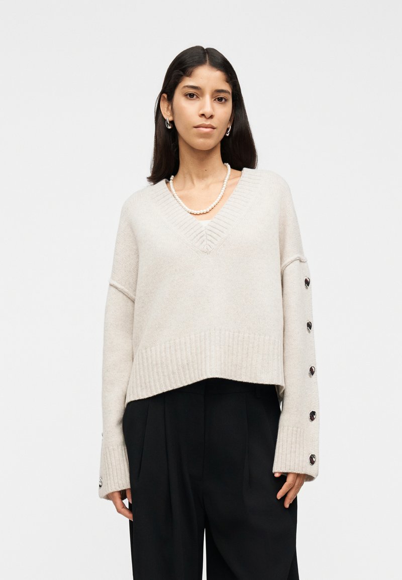 Femme portant un pull à col en V beige avec des boutons sur les manches, un pantalon noir, un collier de perles et des boucles d'oreilles argentées en forme de créole, sur un fond blanc.
