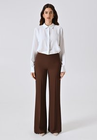 Blusa bianca a maniche lunghe con chiusura a bottoni, abbinata a pantaloni larghi marroni a vita alta, con una texture liscia e linee pulite.
