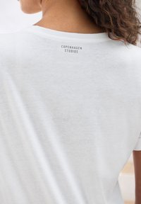 Weißes Baumwoll-T-Shirt mit glatter Textur, mit Rundhalsausschnitt und dem aufgedruckten Text "COPENHAGEN STUDIOS" nahe dem Ausschnitt.